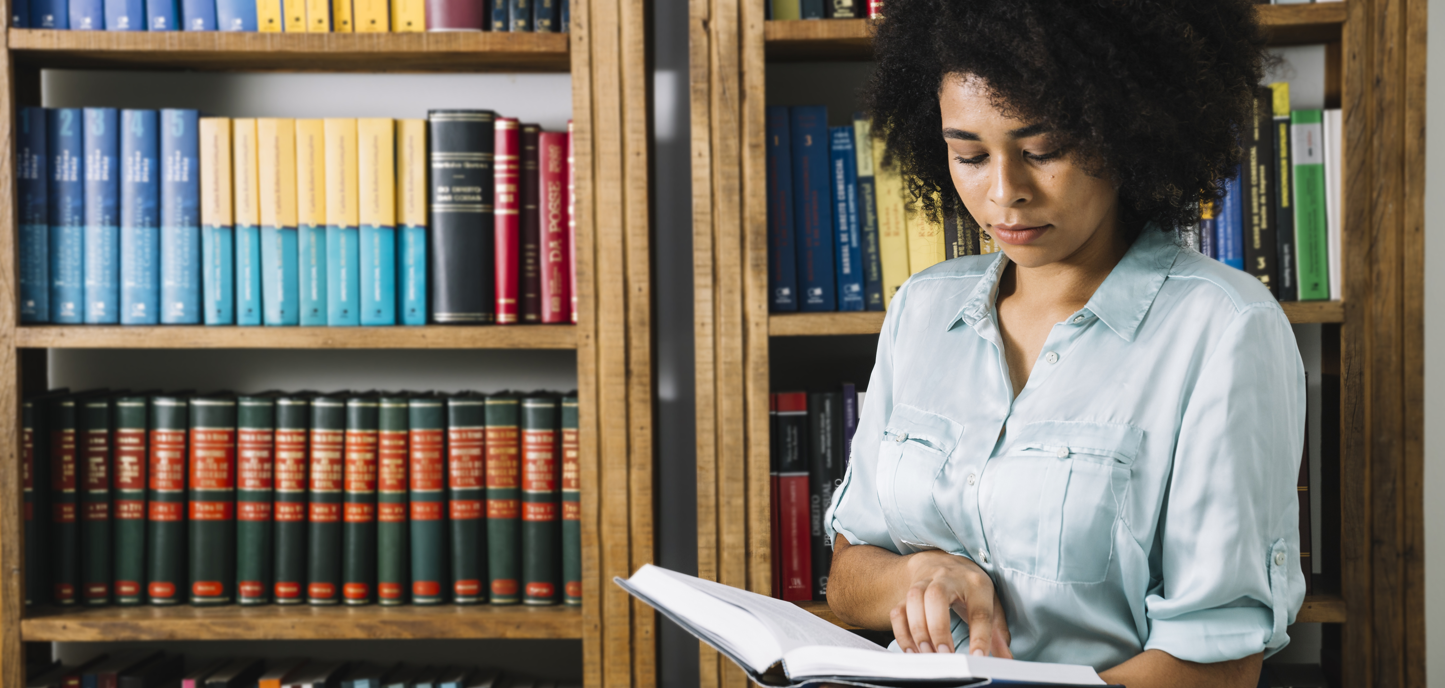 Mulher negra em pé lendo um livro. Ao fundo uma estante de livros